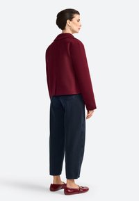 Veste en mélange de laine bordeaux avec une fente dans le dos, assortie à un jean droit bleu marine et des ballerines bordeaux. Texture lisse et design de col classique.