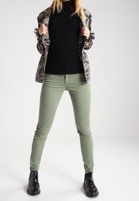 Olivgröna skinny jeans, kombinerade med en svart ribbad topp och en mönstrad camojacka. Inkluderar glansiga svarta ankelskor.