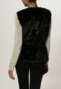 Musta tekoturkisliivi, jossa on korkea kaulus ja takana solmittava yksityiskohta, yhdistettynä vaalean hopeanväriseen, teksturoituun pitkähihainen puseroon. Asun täydentävät mustat housut.