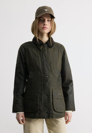 Persoană tânără purtând o jachetă Barbour verde închis și o șapcă asortată, stând pe un fundal simplu deschis, cu brațele relaxate pe lângă corp.