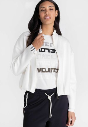 Woman wearing a white zip-up jacket over a printed white shirt and black drawstring pants, posing against a plain white background.