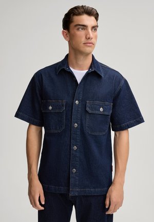 Korte mouwen, donkerdenim shirt met knoopsluiting, twee borstzakken, zilveren hardware en een rechte zoom. Draagt boven een wit shirt.