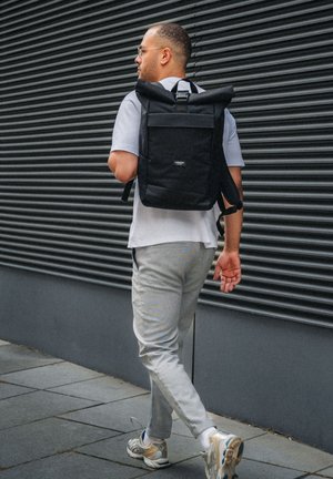 Man som går bredvid en mörk korrugerad vägg, klädd i grå byxor, sneakers, glasögon, en ljusgrå skjorta och en svart rulltoppsryggsäck.