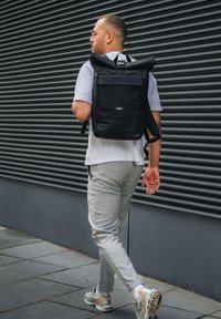 Un homme marchant à côté d'un mur ondulé sombre, portant un pantalon gris, des baskets, des lunettes, une chemise gris clair et un sac à dos noir à enroulement.