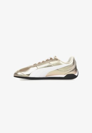 Zapatilla metálica dorada de corte bajo con franja lateral blanca, cordones blancos, cuello acolchado y suela negra, mostrada de perfil sobre fondo blanco.