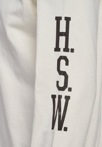 Camisa blanca de manga larga con letras negras en negrita "H.S.W." verticalmente en el lado izquierdo, hecha de suave tela de algodón.