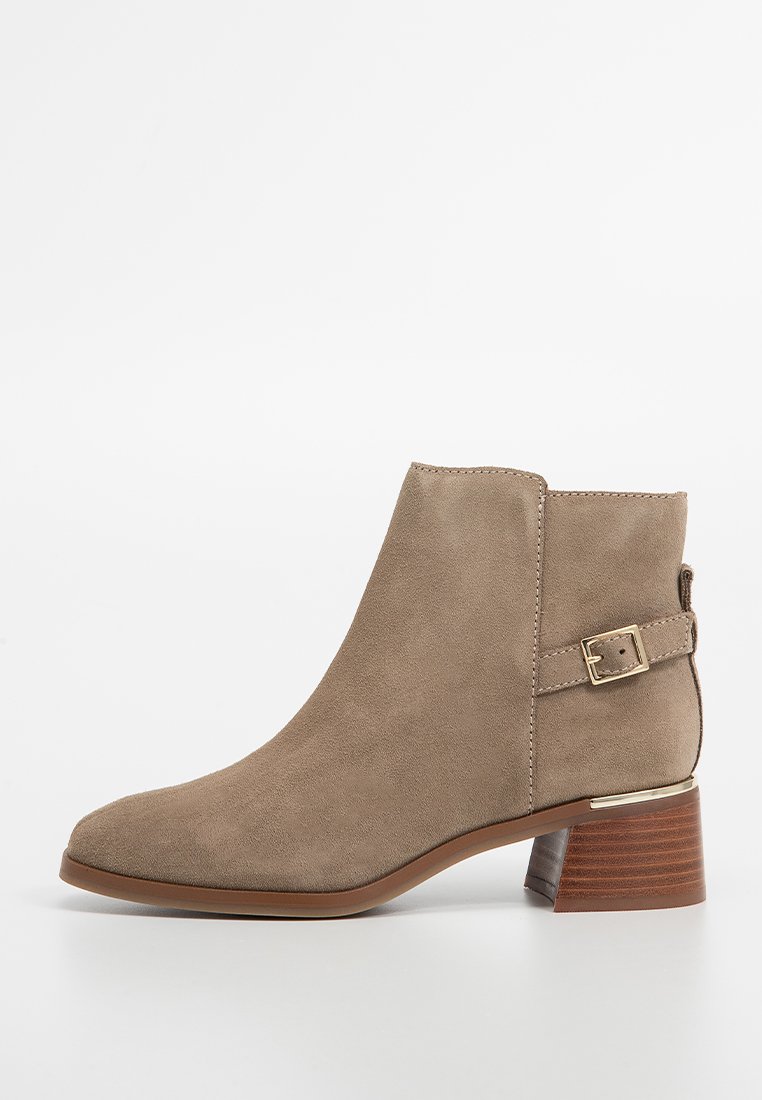 Botte en suede beige à bout rond, avec un accent de boucle en métal doré et un talon en bois brun de hauteur moyenne. Texture lisse.
