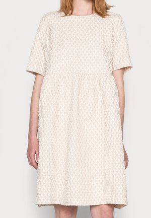 Kobieta w kremowej sukience do kolan, z krótkimi rękawami, o fakturowanej powierzchni, luźnym kroju i okrągłym dekoltem.