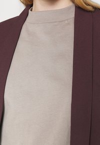 Vaalea beige korkeakauluksinen paita tumman viininpunaisen bleiserin alla, näkyvissä ylävartalo, osa kaulasta ja hiuksista.