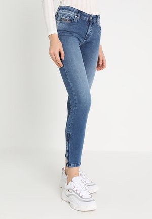 Kobieta w dopasowanych niebieskich dżinsach z bocznymi zamkami błyskawicznymi, zestawiona z białymi masywnymi sneakersami i białym prążkowanym topem z długim rękawem.