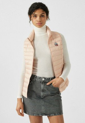 Light pink puffer vest with horizontal quilted design, zip front, and logo patch. Paired with a white ribbed turtleneck and grey denim skirt.