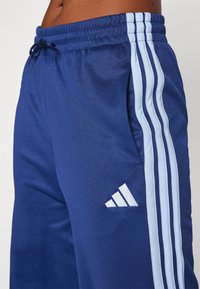 Marine athletic bukser med lyseblå tre-stripes design på siden, justerbar talje, og broderet logo i hvid. Blødt stoftekstur.