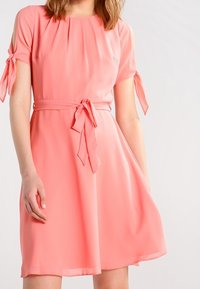 Kortärmad, korallfärgad klänning i lätt tyg med figursydd midja och knytningar på ärmarna. A-linjeformad med en fladdrande kjol.
