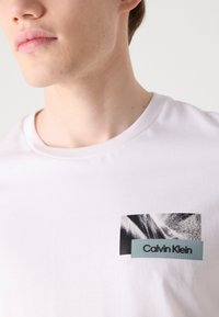 Baltas kokvilnas T-krekls ar grafisku plāksteri melnbaltā dizainā un zilu "Calvin Klein" etiķeti krūšu kabatas zonā.