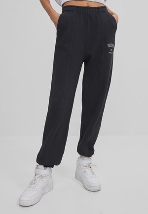 Personne portant un pantalon de jogging noir Yosemite et des baskets montantes blanches, debout les mains dans les poches sur un fond gris.