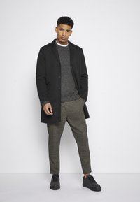 Abrigo negro sobre un suéter gris texturizado, combinado con pantalones de cuadros marrones y zapatillas negras, todo sobre un fondo blanco liso.