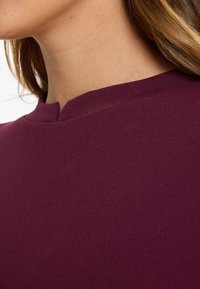 Close-up of a woman's neck and shoulder wearing a maroon top with a small slit notch collar, hair falling behind the shoulder.