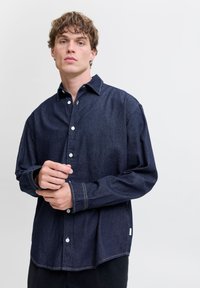 Mørk denimskjorte med spiss krage, hvite knapper, løs passform og kontrasterende sømmer. Har en buet nederkant.