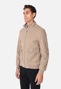 Giacca di lana beige con colletto alto, zip frontale e due grandi tasche anteriori. Polsini e orlo a coste, texture liscia, vestibilità sartoriale.