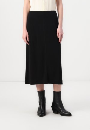 Woman wearing a black midi skirt, ivory sleeveless top, and black leather ankle boots standing against a plain white background.
