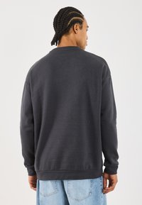 Mann med flettet hår som har på seg en mørk grå genser og lyse blå jeans, står vendt bort fra kameraet mot en hvit bakgrunn.