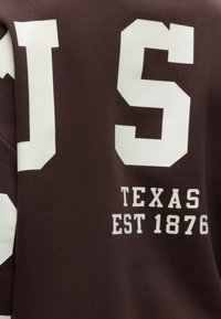 Tissu marron avec de grandes lettres blanches et le texte "TEXAS EST 1876" imprimé dessus.
