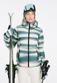 Gestreifte Jacke in Grün- und Cremefarben, mit Kapuze und Reißverschlüssen. Skistöcke und Skier mit Glitzerakzenten in den Händen gehalten.