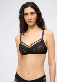 Νεαρή γυναίκα με μακριά σγουρά μαλλιά, που φοράει μαύρο διάφανο σουτιέν από δίχτυ και ταιριαστό μαύρο εσώρουχο, με φόντο απλό φωτεινό.