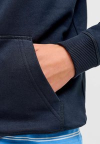 Sweat-shirt bleu marine avec poignet côtelé et poche. Texture lisse avec un détail de couture latérale. Le chemisier en dessous présente des rayures bleues et blanches.