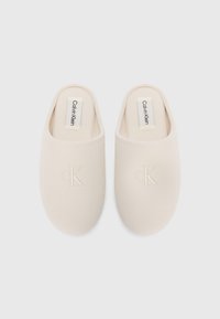 Beige instapmules met een zachte textuur, voorzien van een geborduurd 'CK'-logo aan de voorkant en stoffen labels met 'Calvin Klein'-branding.