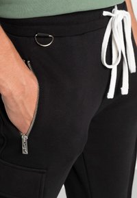 Zwarte sweatpants hebben een met rits afsluitbare zak met zilverkleurige hardware, witte trekkoorden en een textuur van de tailleband. Versierd met een lusdetail.