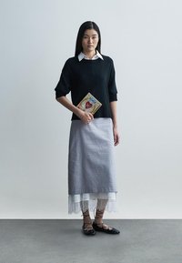 Fekete rövid ujjú kötött pulóver, alatta fehér galléros ing, szürke, csipkével díszített szoknya. A modell egy díszes könyvet tart, fekete lapos cipőt visel.