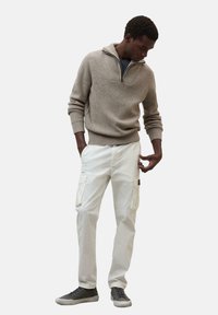 Beige gerippter Pullover mit einem halben Reißverschlusskragen, kombiniert mit weißen Cargohosen mit Taschen. Das Model trägt graue Sneaker.