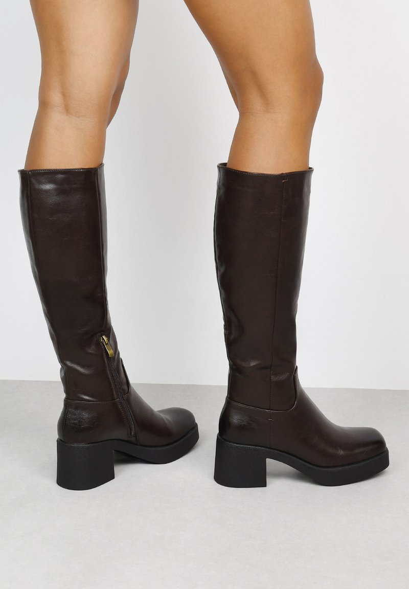 Botas de cuero marrón hasta la rodilla con un acabado texturizado, punta cuadrada y tacón grueso negro. Presentan una cremallera lateral para ponerlas y quitarlas con facilidad.