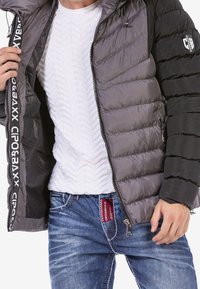 Chaqueta acolchada en negro y gris, con cierre de cremallera y acentos de logo. Camisa blanca texturizada debajo y jeans azules con etiqueta roja.