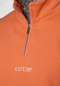 Oranger Pullover mit einem Viertelreißverschluss, aus weichem Material, mit einem weißen "ESTEEM"-Logo auf der Brust und strukturierten Kragenaufsätzen.
