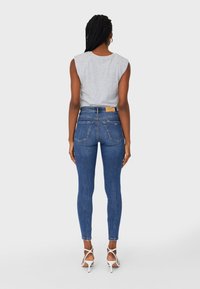 Mujer con trenzas, vistiendo una blusa sin mangas gris, jeans ajustados de tiro alto azules y sandalias de tacón alto blancas, de pie de espaldas.