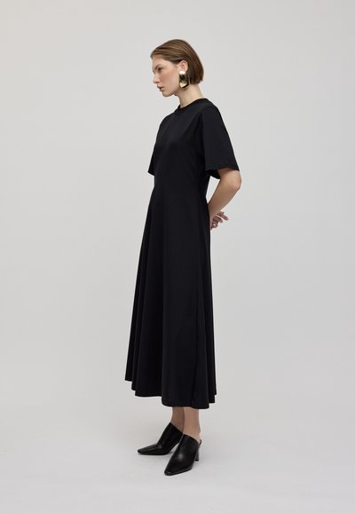Abito nero al ginocchio con maniche corte, realizzato in tessuto morbido, caratterizzato da una vestibilità rilassata e una silhouette fluida. Abbinato a mule neri.