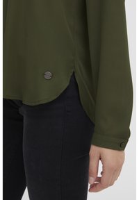 Blusa verde oliva con maniche lunghe; orlo arrotondato con spacchi laterali; presenta un piccolo dettaglio con bottone nero vicino all'orlo.