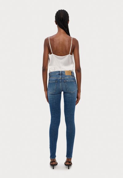 Sötét kék farmer skinny farmerek, klasszikus öt zsebes dizájnnal és barna bőr címkével a hátsó derékszíjon. Fényes fehér szatén felső.