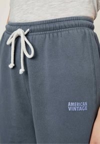 Harmaat collegehousut pehmeästä kankaasta, jossa on joustava vyötärö, valkoinen nyöri ja painettu "AMERICAN VINTAGE" -logo vaaleanharmaana.