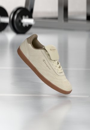 Scarpa da ginnastica Champion beige e marrone che fluttua sopra un pavimento da palestra con manubri neri sfocati sullo sfondo.