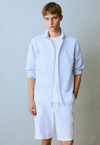 Svetlo modra prugasta srajca s gumbi preko bele majice, v kombinaciji z belimi cargo kratkimi hlačami z stranskimi žepi. Priložnostna in sproščena zasnova.