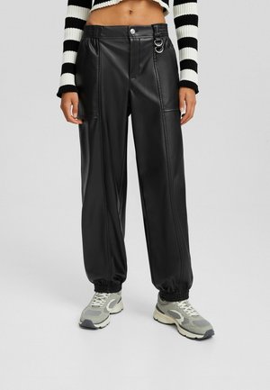 Pantalon de jogging en simili cuir noir avec poignets élastiques, poches avant, doubles anneaux en métal, porté avec des baskets blanches et grises et un pull court à rayures.