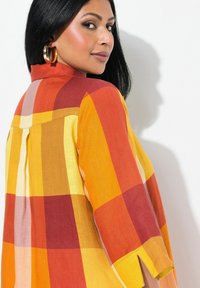 Donna con capelli neri che indossa grandi orecchini a cerchio dorati e una camicia a quadri gialla, arancione e rossa, guardando oltre la spalla.