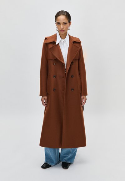 Femme portant un long manteau marron à double boutonnage, une chemise blanche à col, un large jean bleu et des chaussures noires à bout carré, se tenant sur un fond sobre.