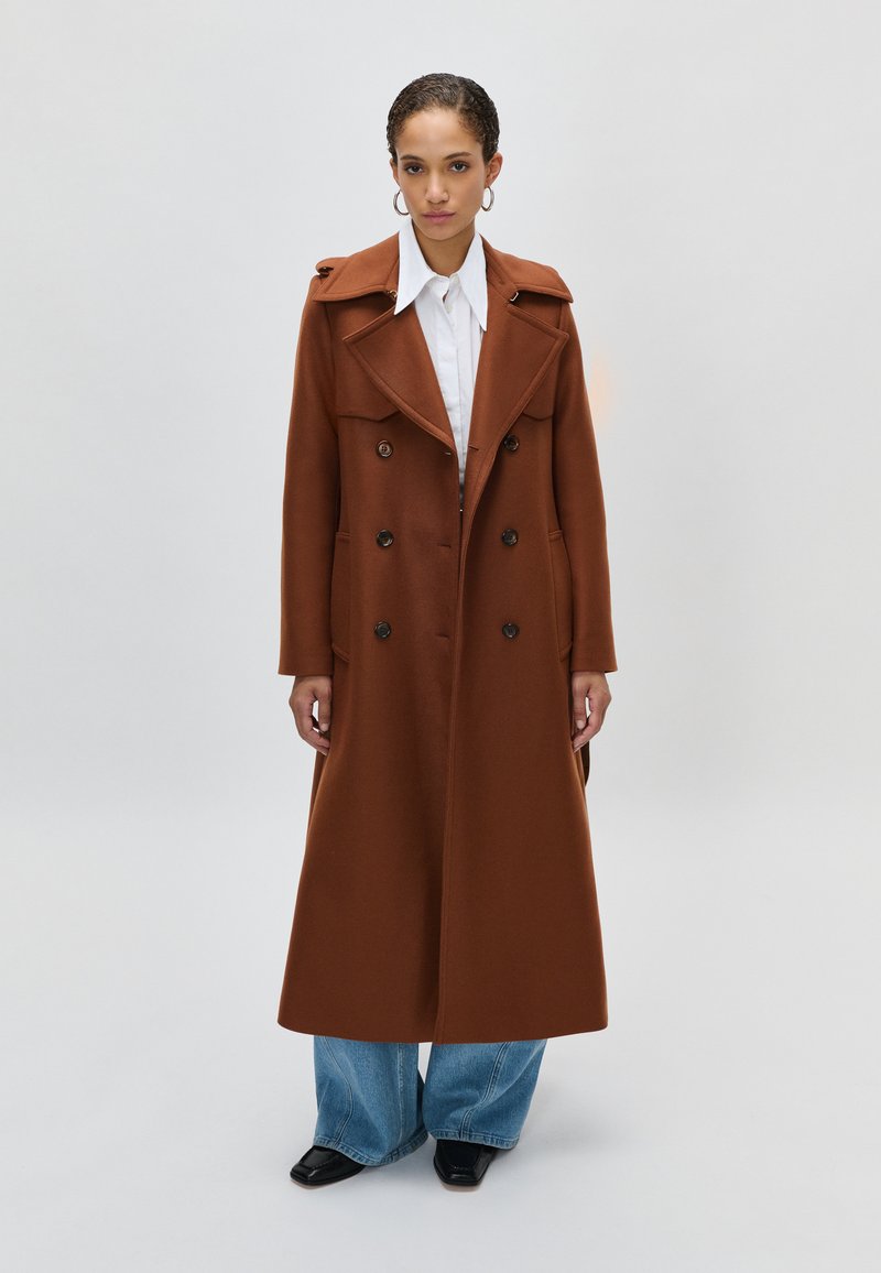 Woman wearing a long brown double-breasted coat, white collared shirt, wide blue jeans, and black square-toe shoes standing on a plain background.