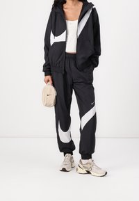 Svart atletisk träningsställ med vita detaljer, elastisk midjereså och jacka med dragkedja. Kombinerat med ljusbeige väska och grå sneakers.