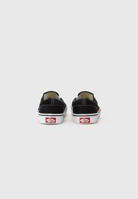 Fekete vászon slip-on cipők fehér gumitalppal, kontrasztos varrással és piros logó részlettel a sarkon. Egyszerű, alacsony profilú dizájn.