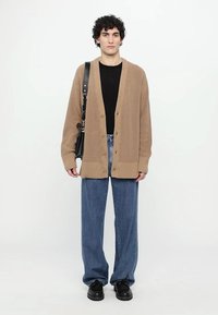 Beige strikket cardigan med knappelukking, brukt over en svart skjorte. Kombinert med løse denimbukser og svarte sko. Minimale tilbehør.
